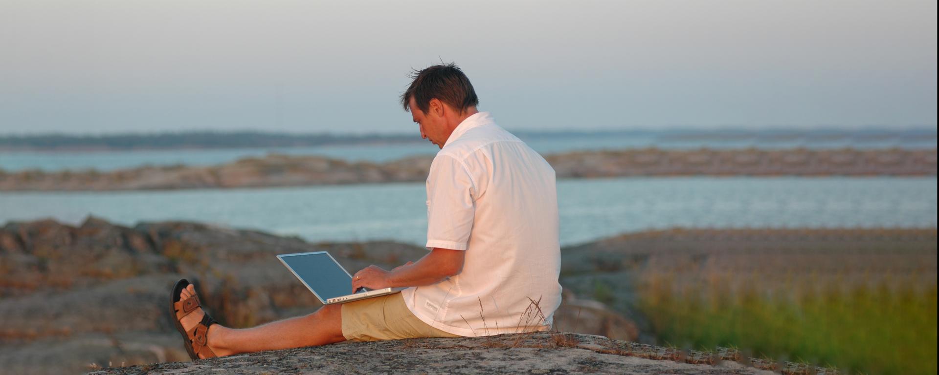 Man med sidan till sitter på klippor vid havet på Åland med en laptop i famnen.